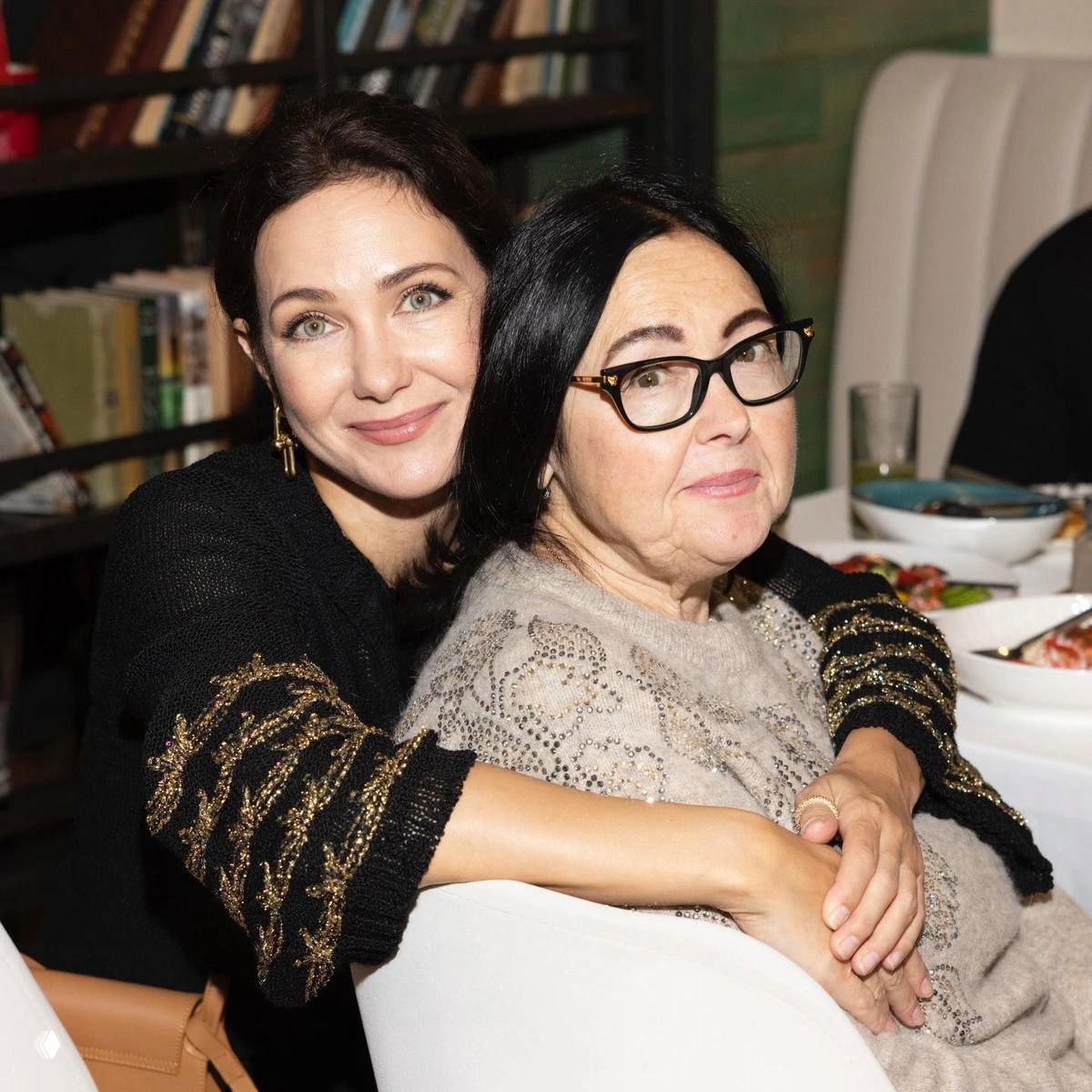 Две женщины сидят вместе за столом в уютной обстановке библиотеки или кафе: объятия и дружеская поддержка, семейная встреча в камерной атмосфере.