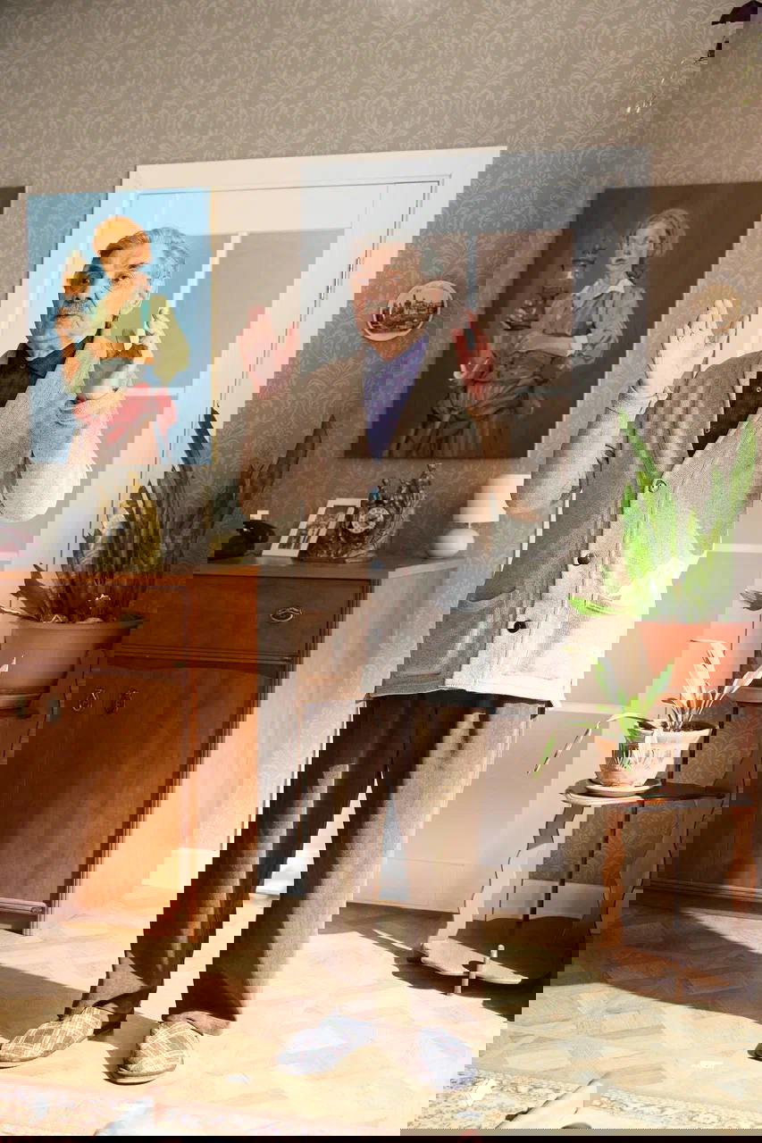 Полноформатный кадр пожилого актёра в жилом интерьере: он стоит перед дверью, жестикулируя в сцене семейного диалога на съёмочной площадке.