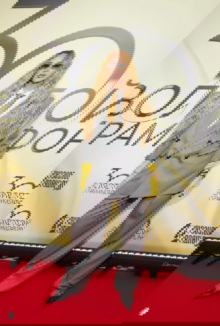 Женщина в свободном светло‑сером костюме, жёлтых перчатках и волнистых волосах позирует на красной дорожке музыкального мероприятия с ярким освещением.