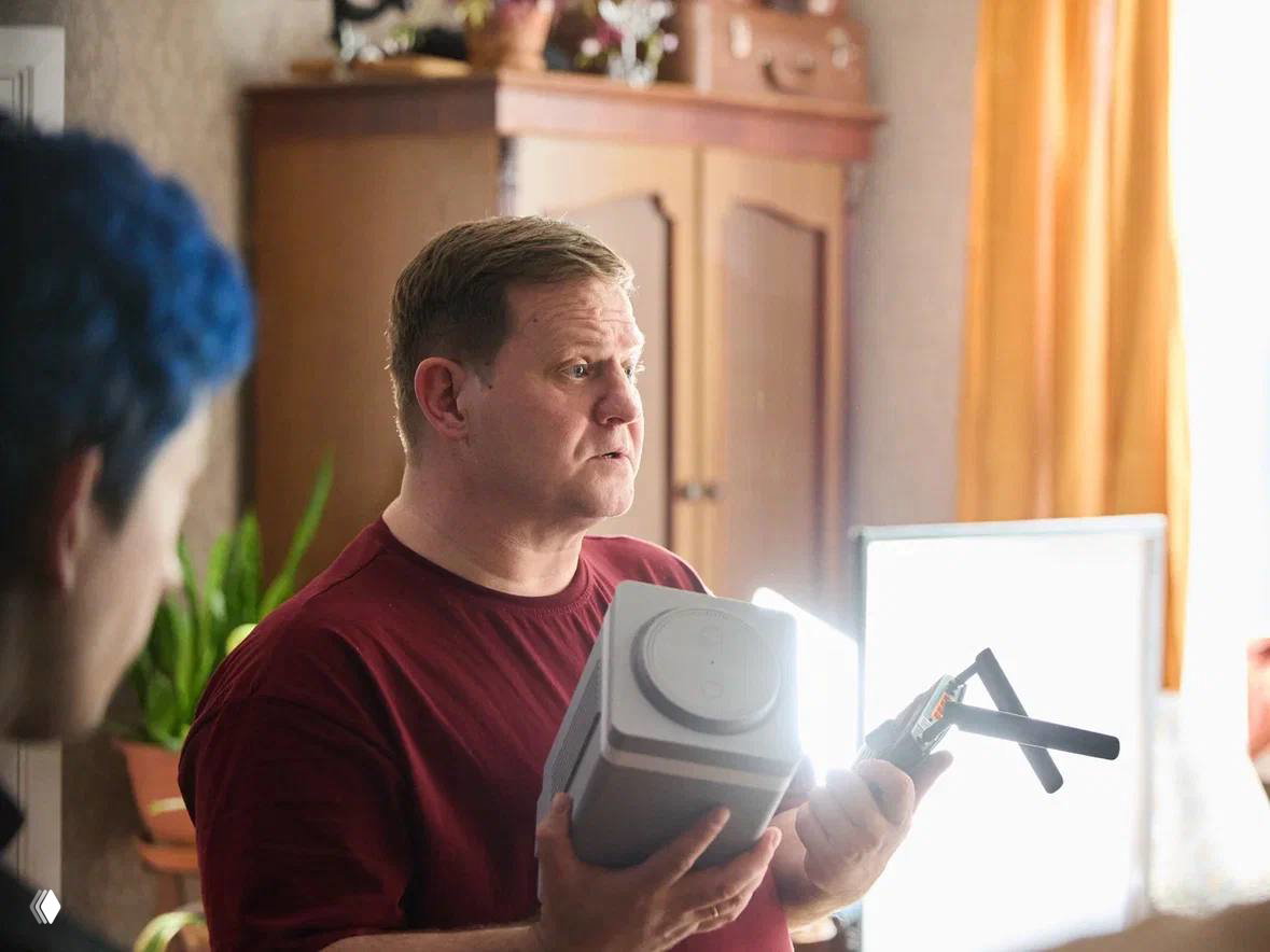 Крупный план актёра в повседневной одежде обсуждающего реквизит: в руках устройство и элементы съёмочного процесса, интерьер комнаты виден на фоне.