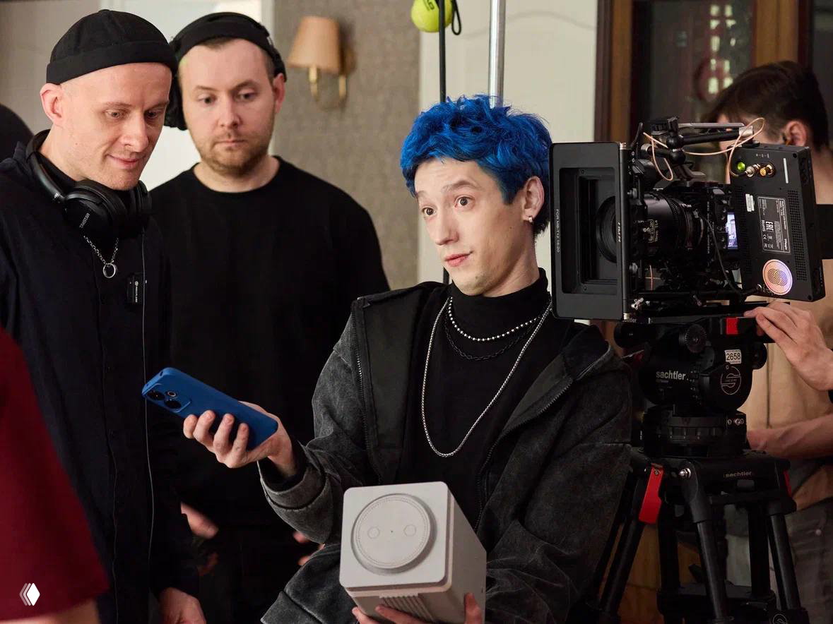 Съёмочная сцена с актёром и членами киносъёмочной группы рядом с камерой: обсуждение кадра и демонстрация смартфона и реквизита.