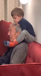 Пожилой мужчина сидит на диване, на его плечах сидит маленький ребёнок; кадр из видео с тёплой семейной атмосферой.
