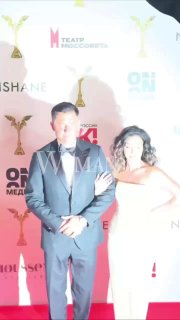 Мужчина и женщина в вечерних нарядах идут по красной дорожке церемонии «Ника», позируют на фоне брендволла с логотипами