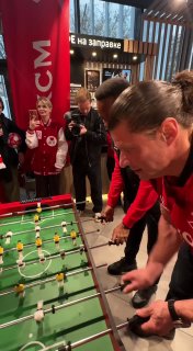Валерий Юшкевич сосредоточенно играет в настольный футбол на фоне логотипов АЗС ЛУКОЙЛ и болельщиков, кадр с мероприятия акции.