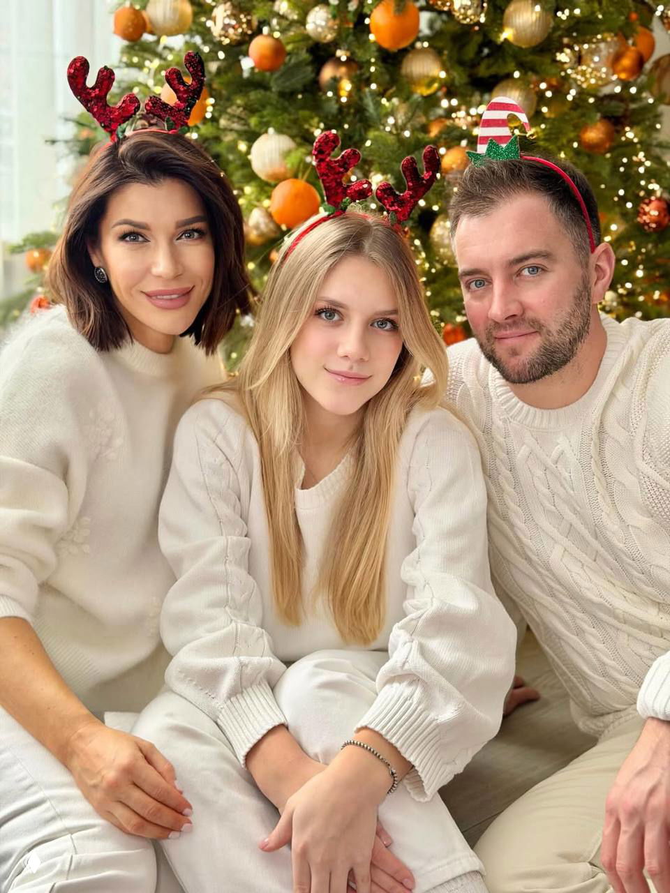 Трое людей в белой и светлой одежде у большой ёлки, у двоих на голове праздничные рожки — тёплая праздничная сцена.