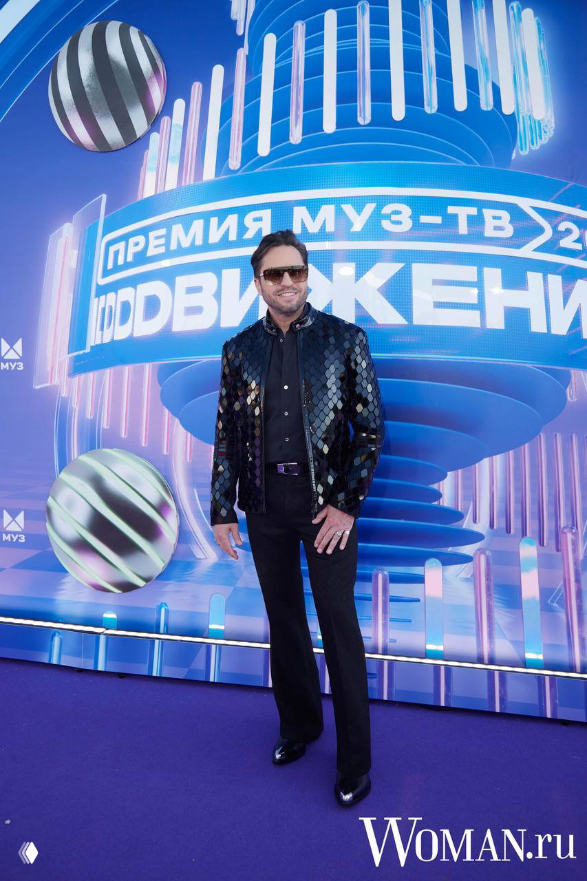 Мужчина в блестящей куртке и тёмных брюках позирует на фиолетовой дорожке премии МУЗ‑ТВ «Движение» с декорациями в виде логотипа.
