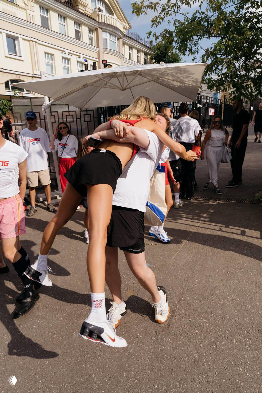 Уличная сцена: гости мероприятия обнимаются и поздравляют друг друга возле зонта и столиков на открытом воздухе в солнечный день.