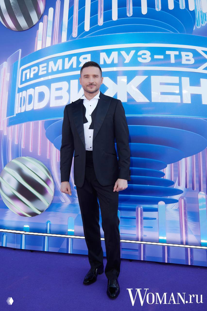 Мужчина в классическом чёрном смокинге с белой рубашкой позирует на фоне сценического оформления премии МУЗ‑ТВ «Движение».