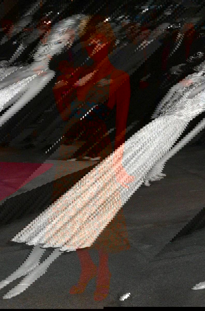 Женщина в светлом летнем платье держит маленькую собачку на светском мероприятии, вокруг люди и охрана, ночная уличная сцена в фоне.