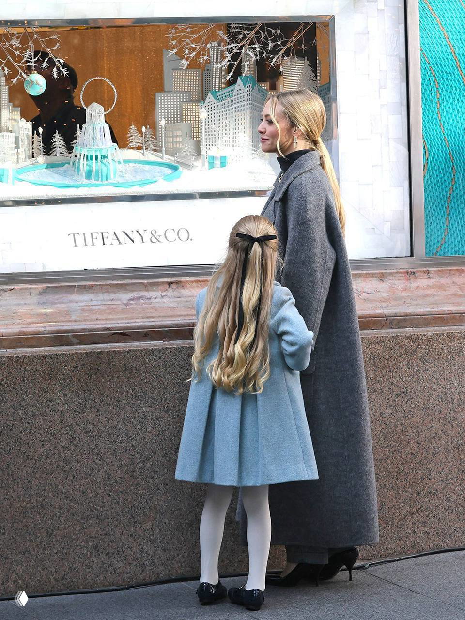 Аманда Сейфрид и девочка рассматривают витрину Tiffany & Co.; рождественская витрина с украшениями и декором на заднем плане.
