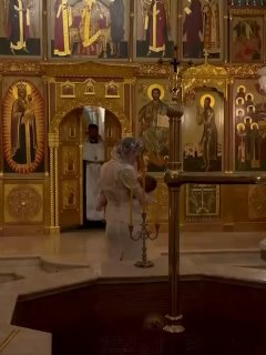 Валентина Иванова в кружевном платке идёт по алтарю храма во время обряда крещения; на заднем плане иконы и церковные свечи.