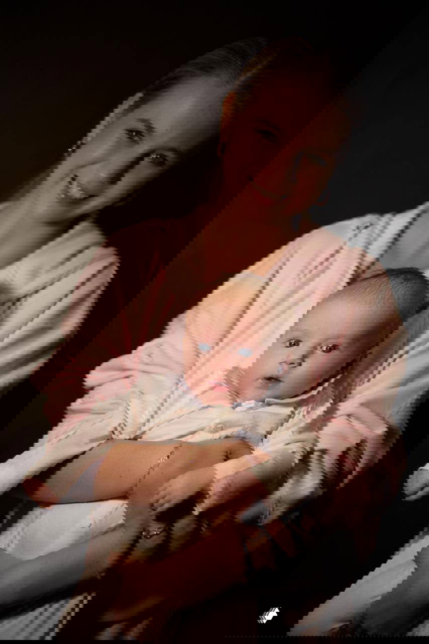 Саша Трусова улыбается и держит на руках новорождённого Мишу в розовой толстовке — полный рост кадра в тёплом освещении.