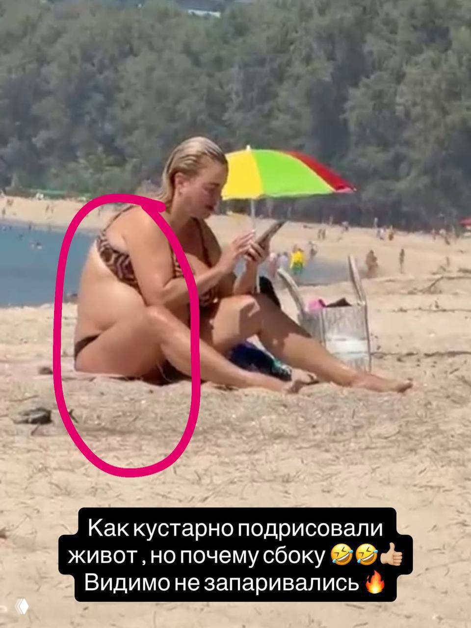 Пляжный снимок: человек в купальнике сидит на песке с телефоном, рядом зонтик и люди на заднем плане.