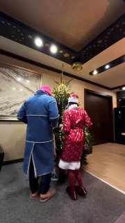 Два человека в костюмах Деда Мороза и Снегурочки сняты сзади в холле отеля — кадр из короткого видео, видны потолочные светильники и ёлка.