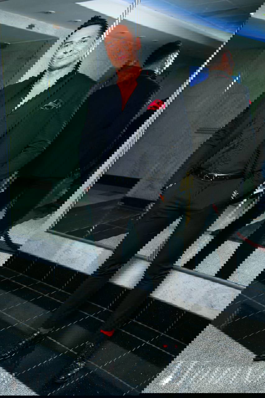 Стас Костюшкин в черном костюме с красным платком в кармане позирует у зеркальной стены в фойе, видно полный рост и отражение.