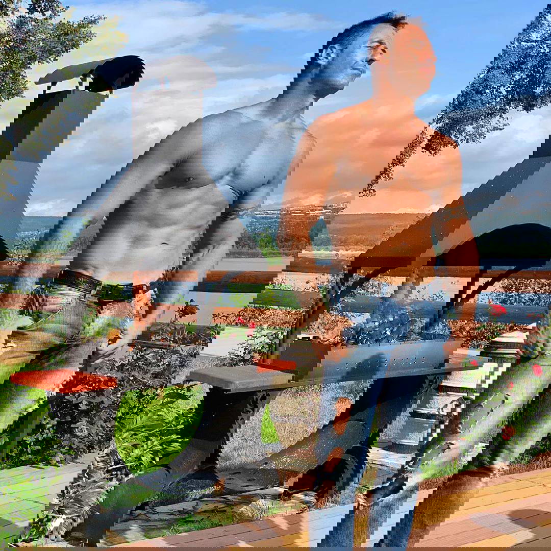 Стас Костюшкин без рубашки у мангала на фоне реки и зелени в рваных джинсах: видна рельефная мускулатура и летний пейзаж на заднем плане.