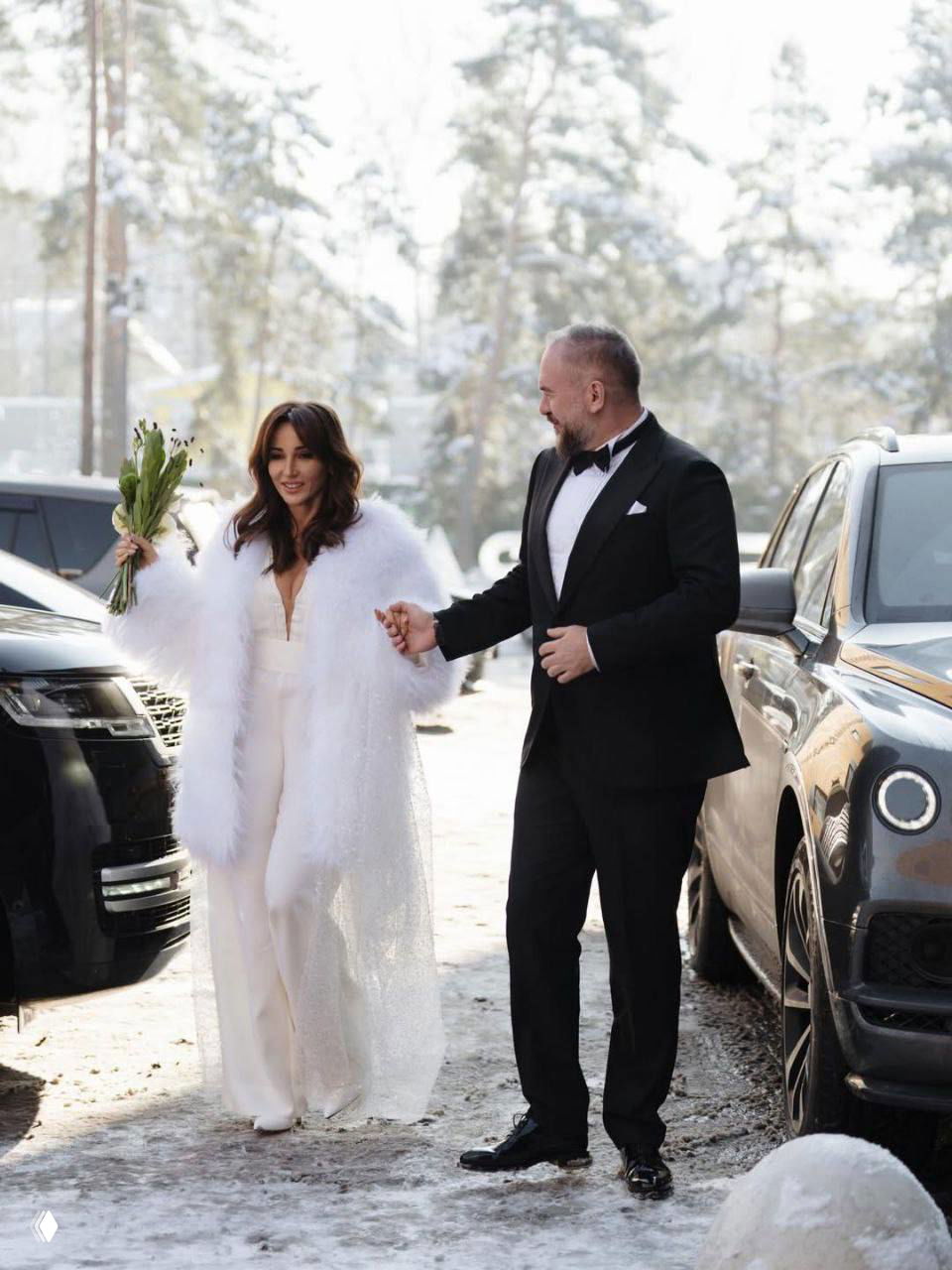 Невеста в белой накидке и жених в смокинге идут по заснеенной улице между автомобилями; невеста держит букет.