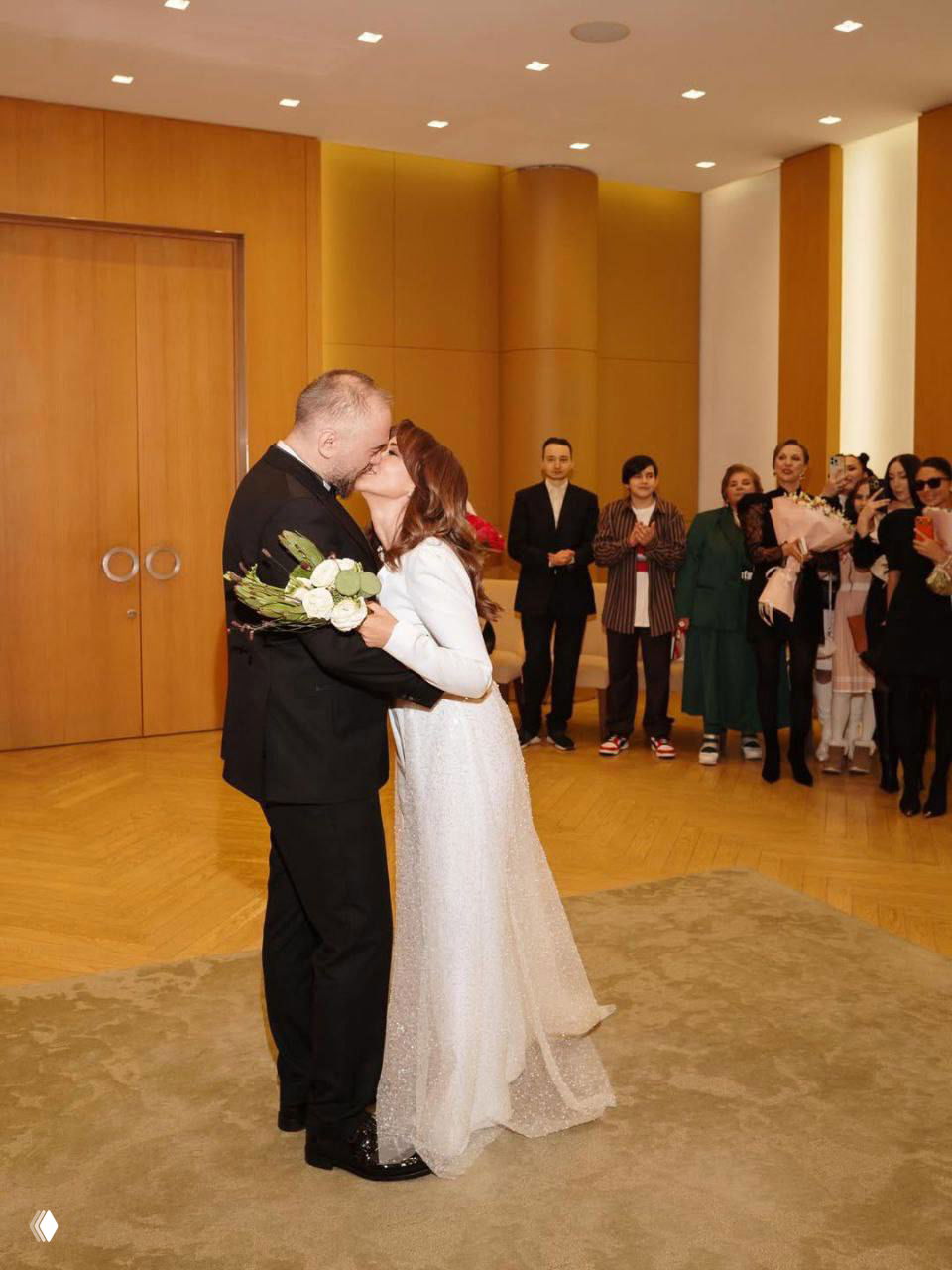 Пара в свадебных нарядах целуется в зале; на заднем плане гости с букетами, невеста держит букет и платье с шлейфом.
