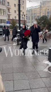 Двое людей с букетами идут по улице возле театра, один прикрывает лицо рукой; на изображении видна урбанистическая сцена и водяной знак WOMAN.ru