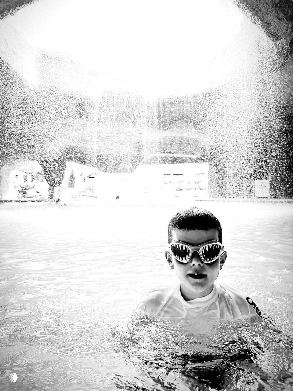 Черно-белое фото ребёнка в бассейне в защитных очках для плавания на фоне падающей воды и купольной зоны аквапарка; семейная сцена отдыха на море.