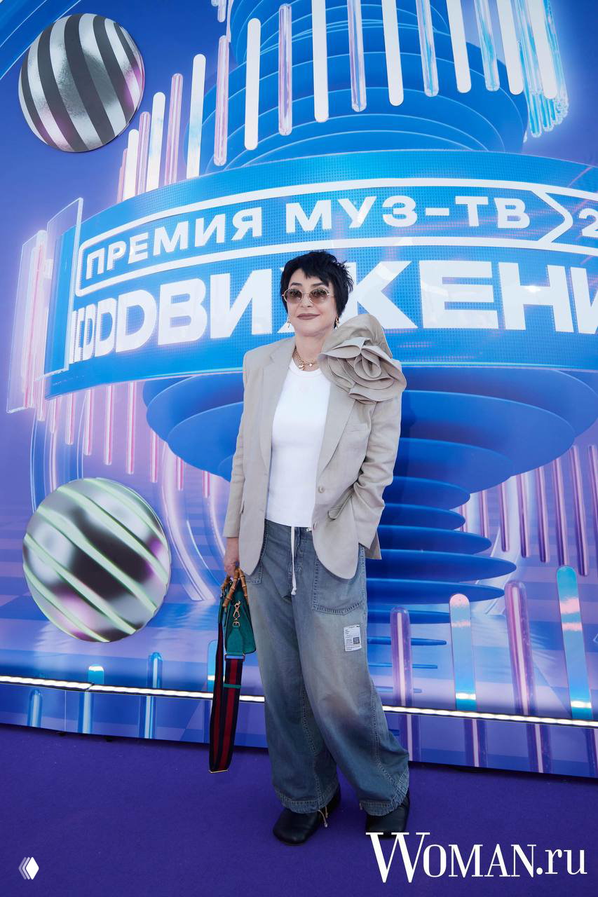 Гостья в светлой жакетной куртке и широких джинсах с крупным декоративным цветком на плече позирует на фоне баннера Премии МУЗ‑ТВ.