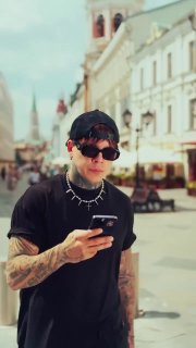 Вертикальный кадр мужчины с татуировками и чёрной одеждой, он держит смартфон на фоне пешеходной улицы и исторических зданий.