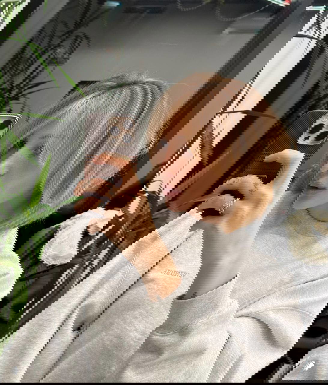 Зеркальное селфи женщины со светло-русой стрижкой каре и чёлкой, держит смартфон в салонной обстановке с растениями на заднем плане