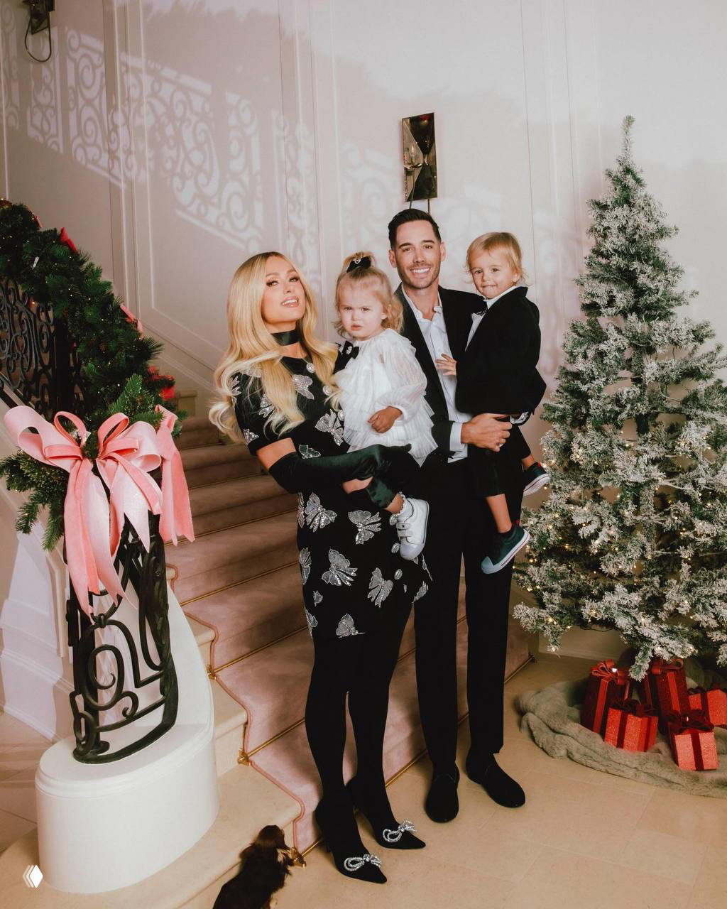 Семейный кадр: женщина и мужчина держат двух маленьких детей у украшенной рождественской ёлки в домашней обстановке, тёплая семейная сцена и интерьер.