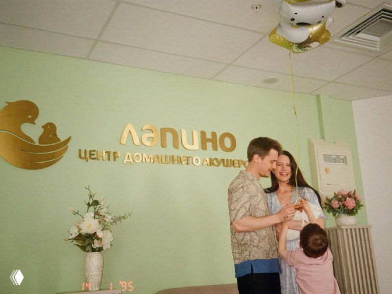 Иван и Диана улыбаются у вывески «Лапино Центр домашнего акушерства», рядом ребёнок и букет — выходной кадр семьи из центра родовспоможения
