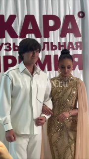 Ольга Бузова и Прохор Шаляпин на фоне баннера ЖАРА Music Awards: пара в вечерних образах позирует перед началом церемонии, водяной знак Woman.ru.