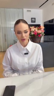 Яна Кошкина в белой блузе сидит за столом во время видеозаписи: видно букет цветов и интерьер жилой комнаты на заднем плане, крупный план.