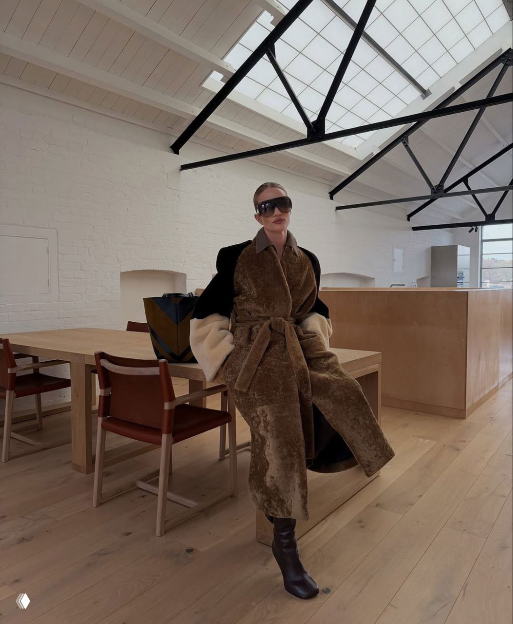 Женщина в длинной коричневой шубе сидит в минималистичном светлом помещении с деревянной мебелью и большими окнами — интерьер и образ.