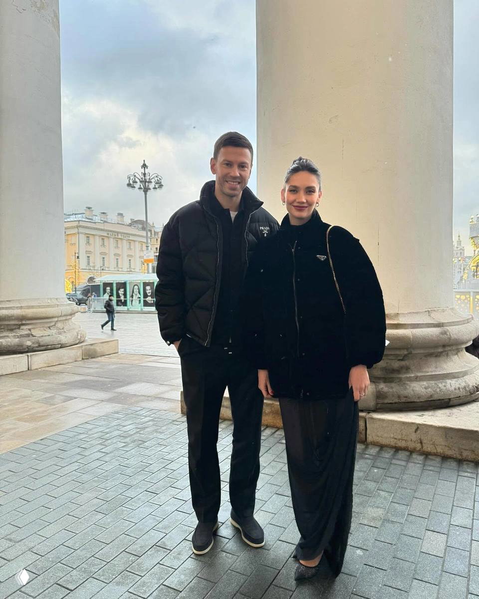 Двое людей в тёплой одежде стоят у колонн на городской площади; на заднем плане видны здания, трамвай и пасмурное небо.