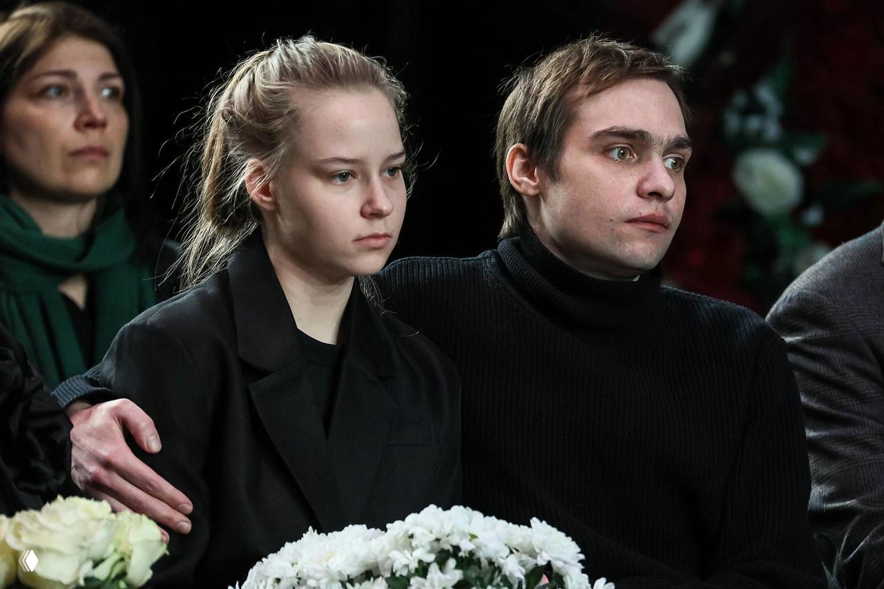 Молодой мужчина и девушка в тёмной одежде сидят рядом на поминальной церемонии, серьёзные лица, рядом лежат букеты и заметна траурная сцена мероприятия.