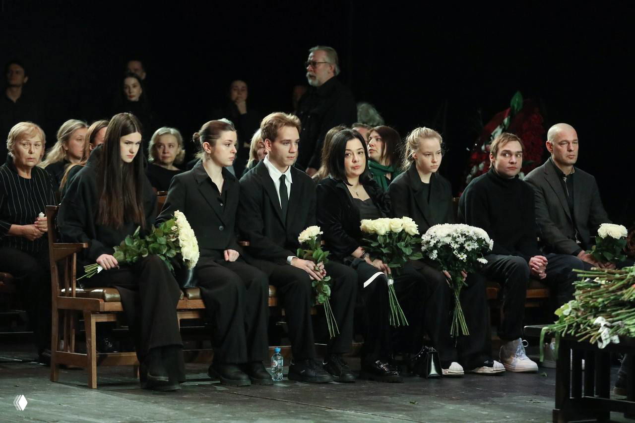 Широкий кадр траурной церемонии в театре: несколько людей в чёрном сидят в ряду и держат букеты белых цветов, приглушённый свет сцены и торжественная атмосфера.