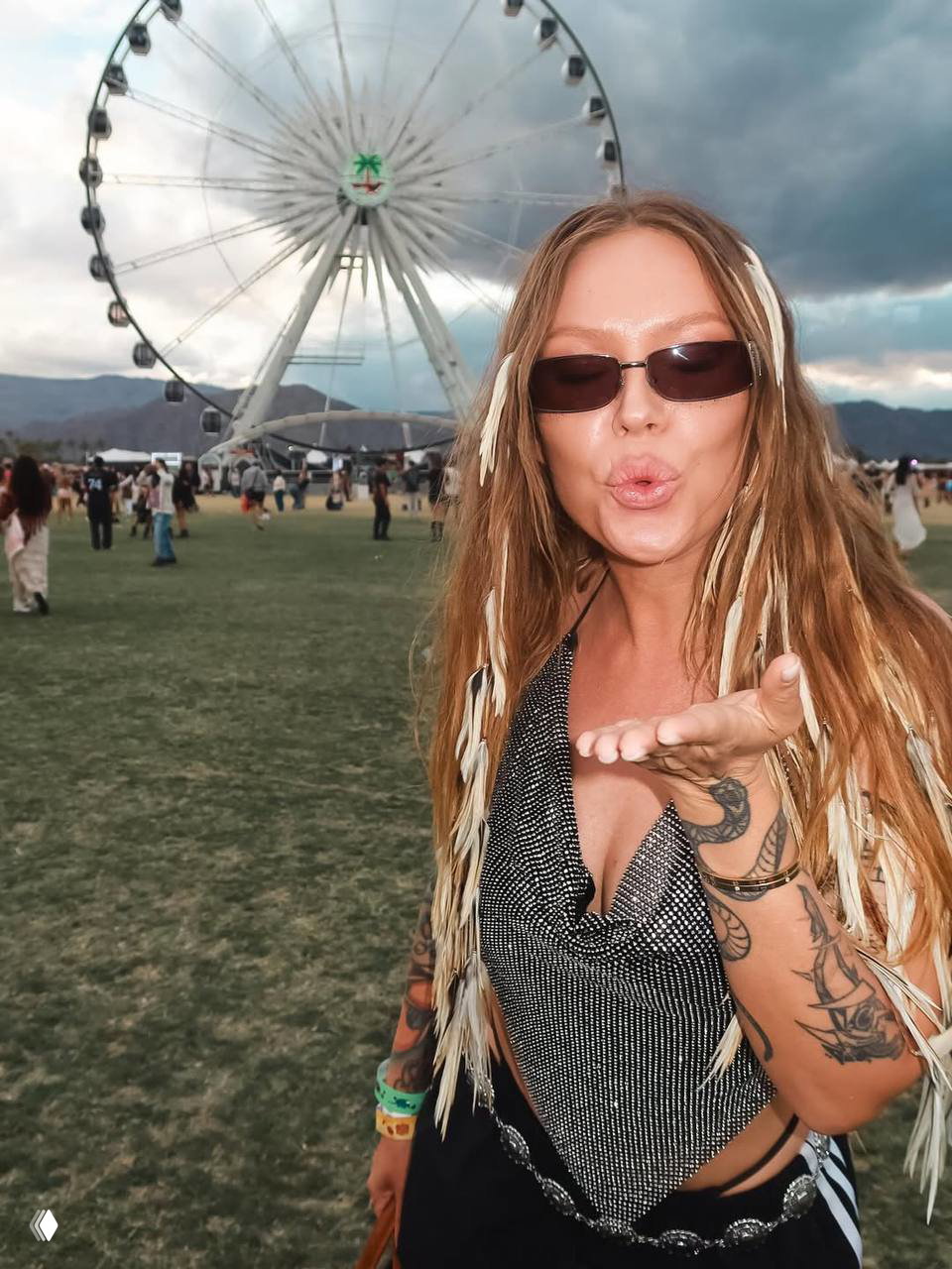 Рита Дакота посылает поцелуй в камеру в серебристом топе и узких солнцезащитных очках на фестивальной поляне Coachella, вокруг — посетители и колесо обозрения.