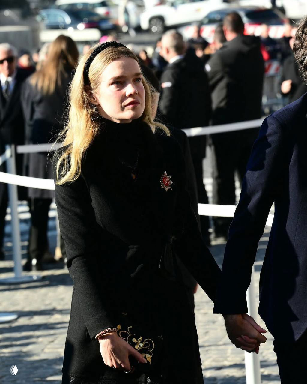 Женщина в чёрном пальто и ободке на голове держит руку другого человека, вокруг ограждения и собравшиеся гости церемонии.