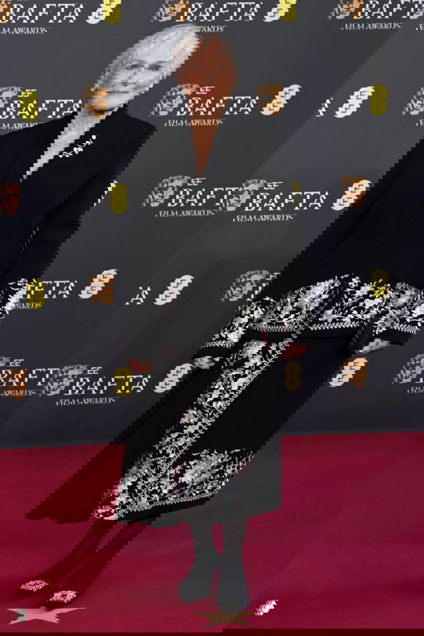 Женщина в тёмном комплекте с декоративной вышивкой и жакетом стоит на красной дорожке BAFTA на фоне пресс-волла с логотипами.