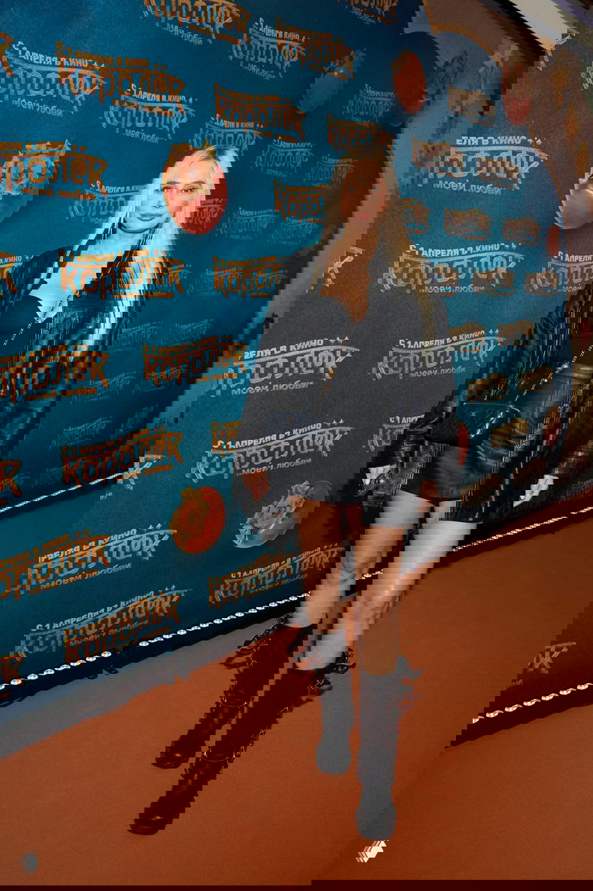 Гостья в чёрном кожаном плаще и мини-юбке стоит у бренд-стены премьеры «Королёк моей любви», видна бренд-оформление и оранжевый ковер.