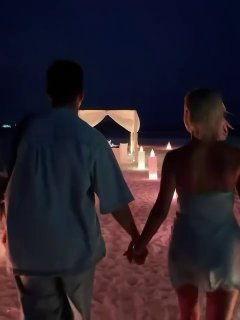 Пара идёт по песчаному пляжу вечером, держась за руки на фоне арки и светящихся украшений — кадр сзади.