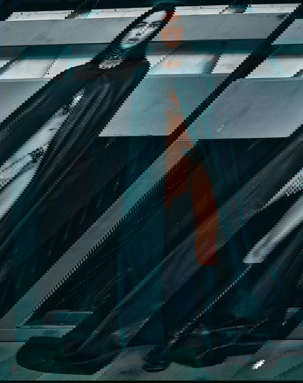 Модель в длинном чёрном плаще и ошейнике стоит в архитектурной студии — минималистичный, строгий кадр с акцентом на силуэт и текстуру кожи.