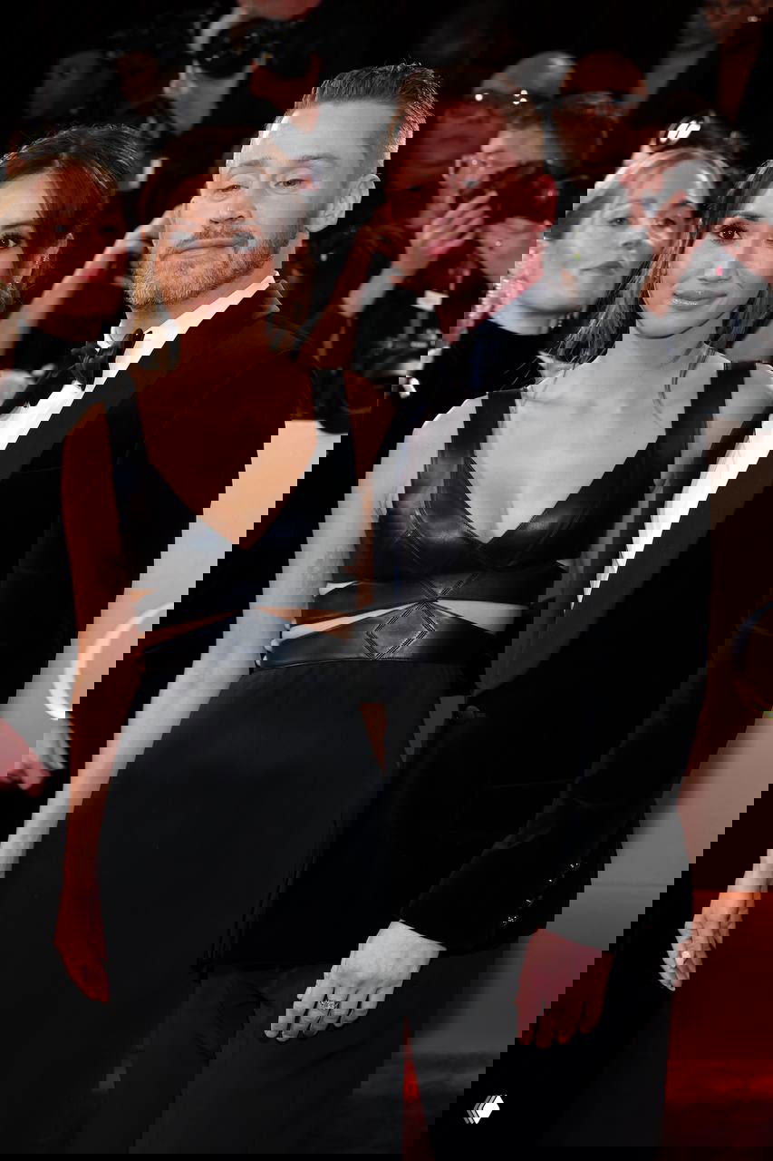 Крупный план пары на красной дорожке: мужчина в смокинге и женщина в чёрном вечернем платье на фоне фотокамер.