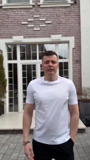 Кадр из видеозаписи: мужчина в белой футболке стоит у фасада здания, на заднем плане видно вход с дверью и окна.