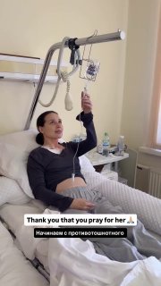 Пациентка сидит на кровати и держит инфузионную стойку, видны капельница и шапочка, рядом тумбочка с бутылкой воды.