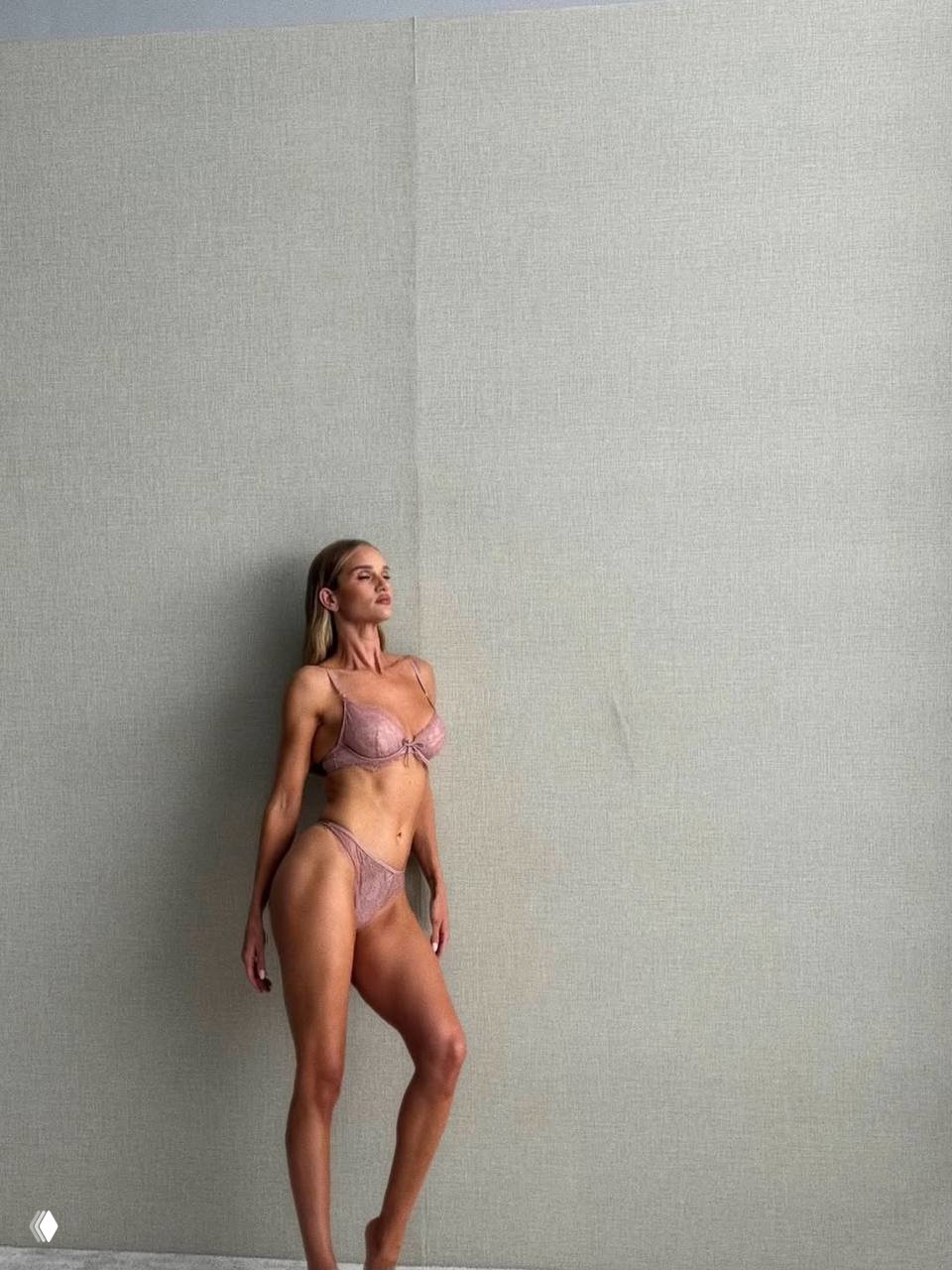 Ростовой кадр Роузи Хантингтон-Уайтли в розовом белье на однотонном фоне; модель стоит, демонстрируя силуэт и фактуру комплекта в студийном освещении.