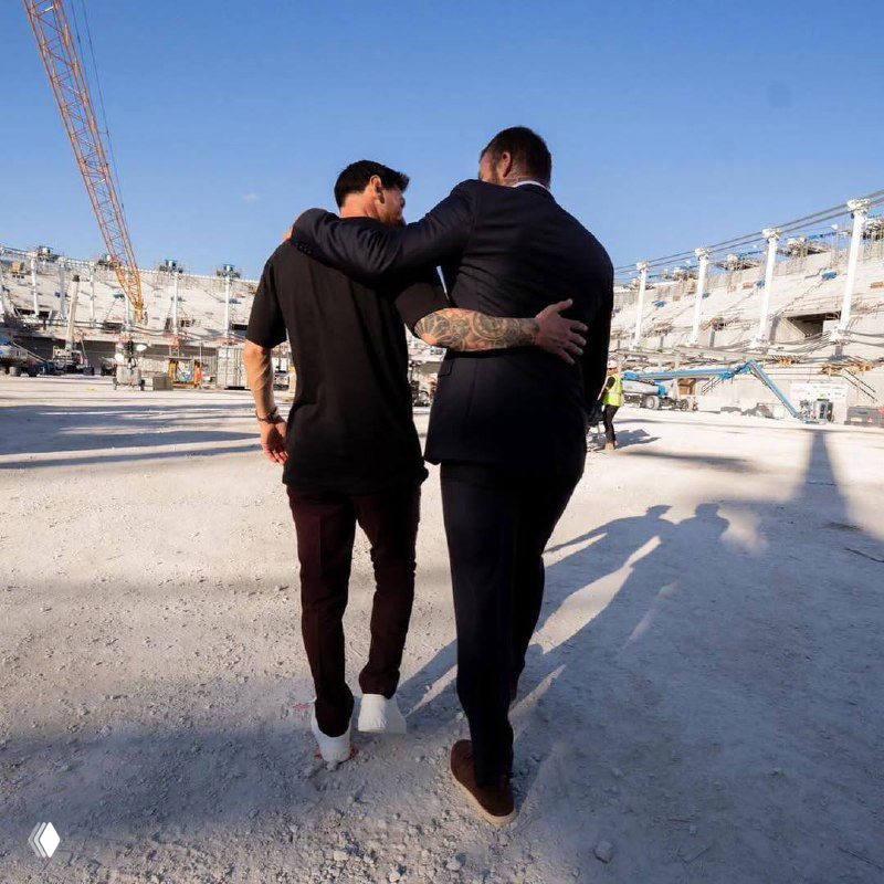 Кадр с рабочей территорией стадиона и проходящими людьми, изображение частично искажено цифровыми артефактами; видна строительная площадка и тени.