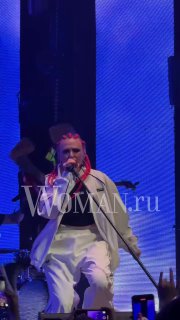 Крупный план солиста группы «Вирус!» на сцене VK Gipsy: микрофон и эмоциональное исполнение, белая одежда и LED‑подсветка, заметен логотип Woman.ru