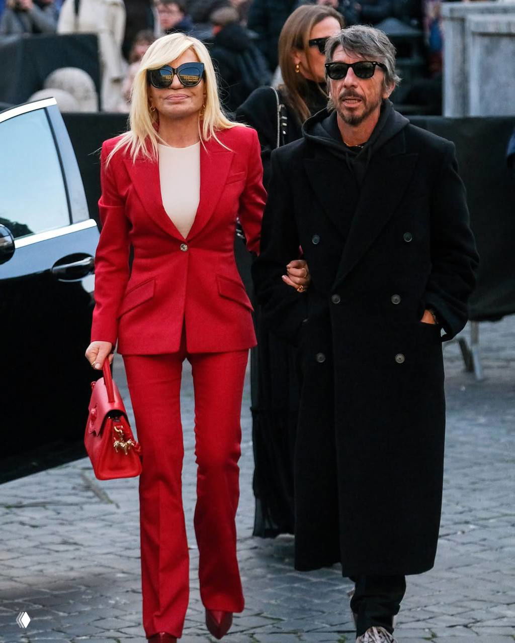 Пара гостей идёт по брусчатке: женщина в ярком красном костюме и сумке, мужчина в тёмном пальто; сцена у траурного мероприятия.