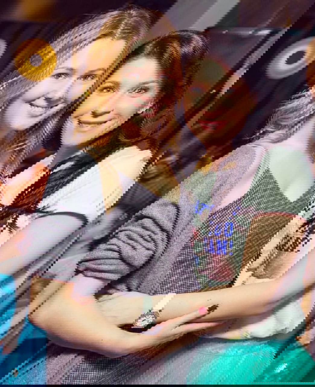 Две улыбающиеся женщины обнимаются на светском мероприятии, фон с приглушённым освещением и гостями в кадре.