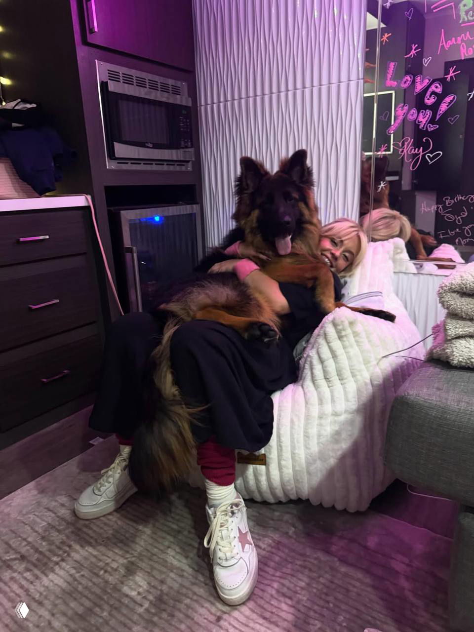 Фото: Сидни Суини держит большую овчарку Салли на коленях в комнате с неоновыми надписями. Видны уши собаки и ноги актрисы.